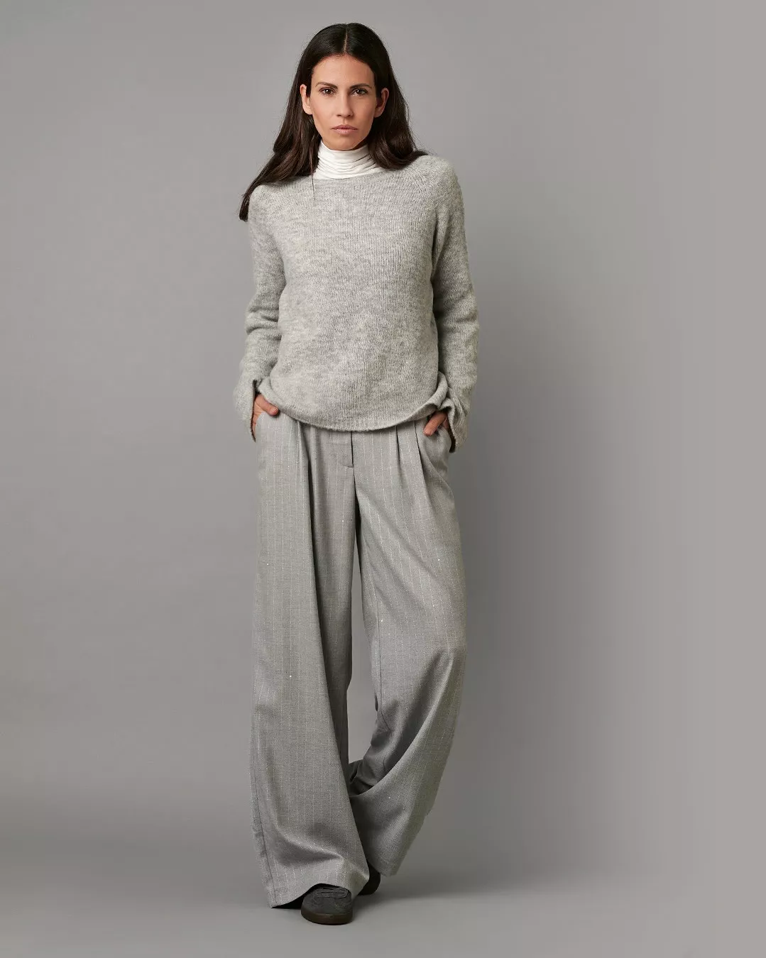 Frau in hellgrauem Strickpullover mit weißem Rollkragen und weiter grauer Stoffhose vor neutralem Hintergrund.