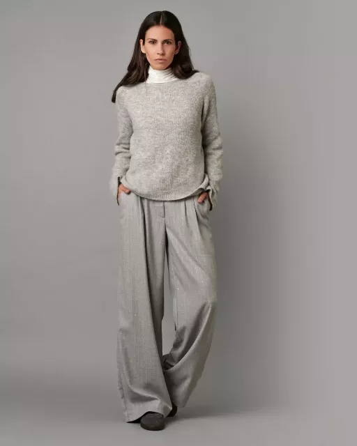 Frau in hellgrauem Strickpullover mit weißem Rollkragen und weiter grauer Stoffhose vor neutralem Hintergrund.